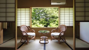 Una habitación japonesa tradicional con suelo de tatami, dos sillas de madera, una mesita y un ventanal con vistas a la vegetación.