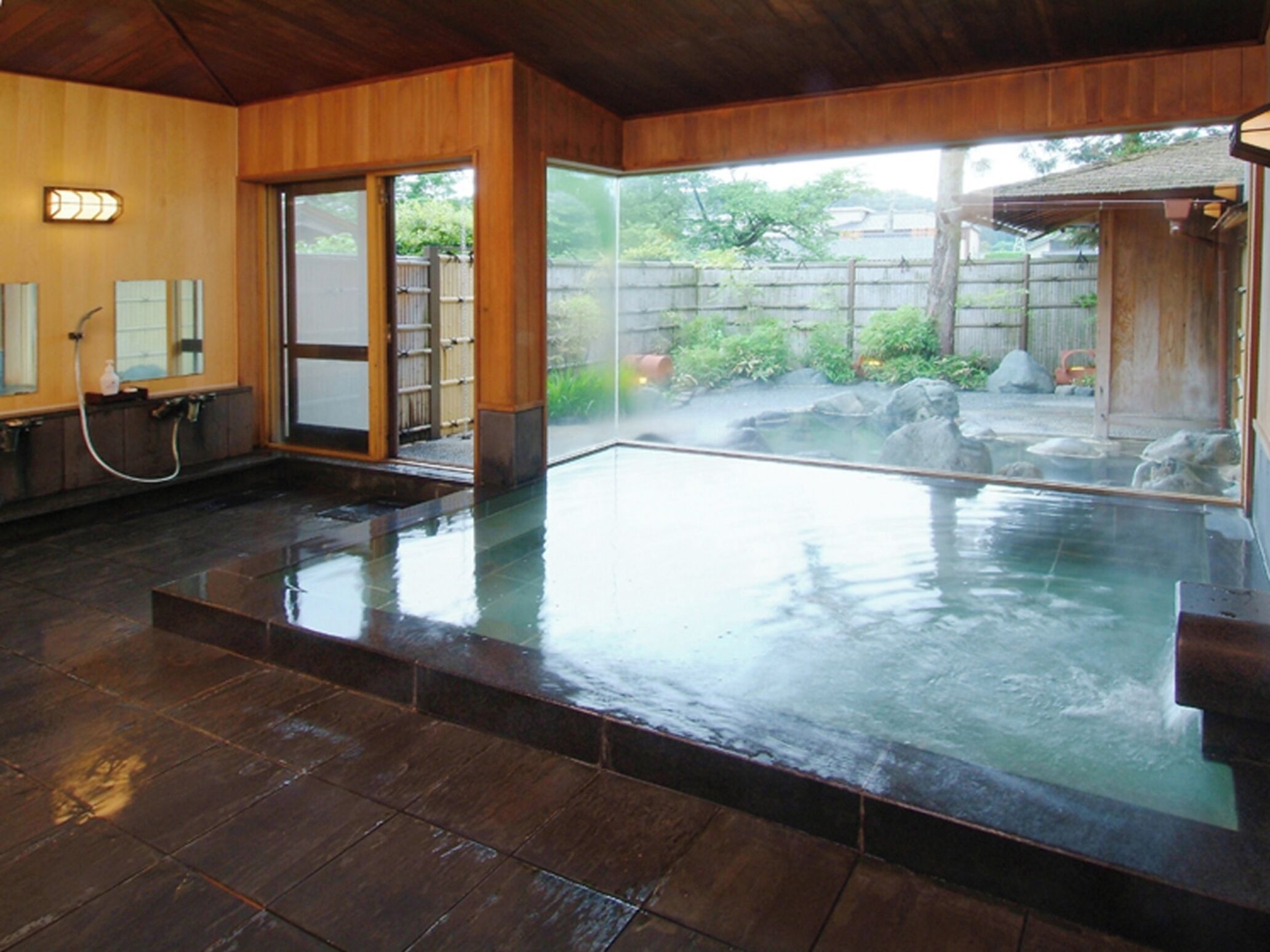A traditional Japanese onsen with a wooden interior, a glass-enclosed hot spring, and a view of the surrounding garden.