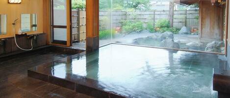 A traditional Japanese onsen with a wooden interior, a glass-enclosed hot spring, and a view of the surrounding garden.