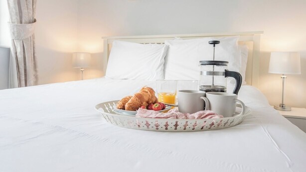 Een opgemaakt bed met een ontbijttafel waarop croissants, aardbeien en sinaasappelsap staan, samen met twee witte mokken en een koffiezetapparaat.