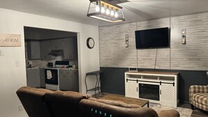 Moderni olohuone, jossa on sohva, seinälle asennettu televisio, takka ja taustalla keittiö.