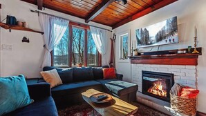 A cozy living room with a fireplace, a large window with curtains, a sofa, and a television mounted above the fireplace.