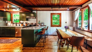 A kitchen with wooden flooring, a dining area with a table and chairs, and a window with a view of greenery.