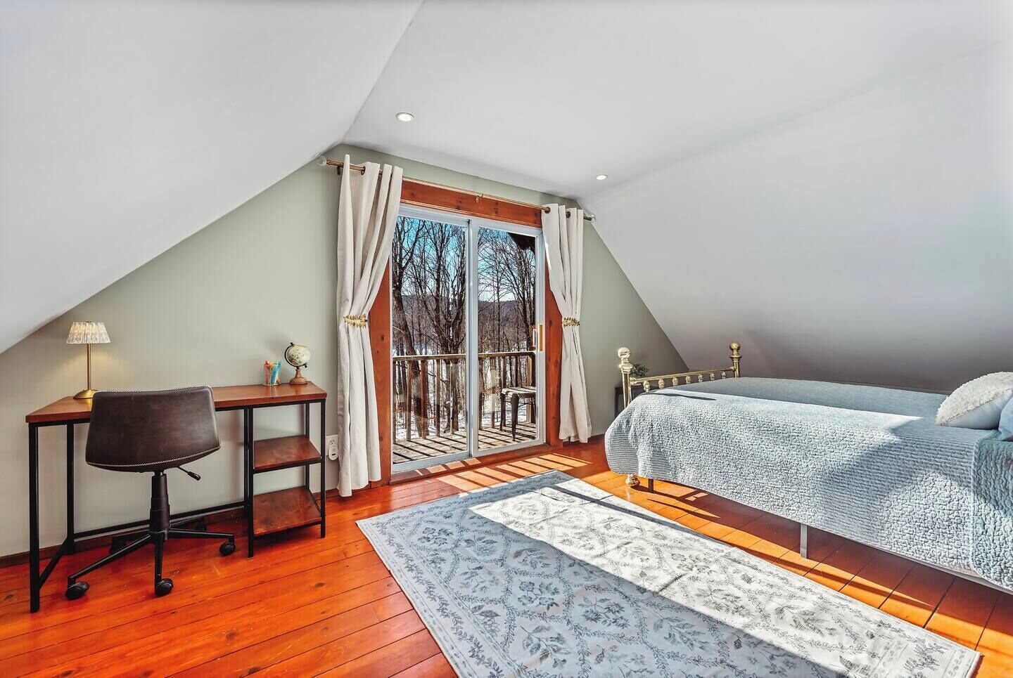 A bedroom with a sloped ceiling, a bed, a desk with a chair, and a large window with curtains.