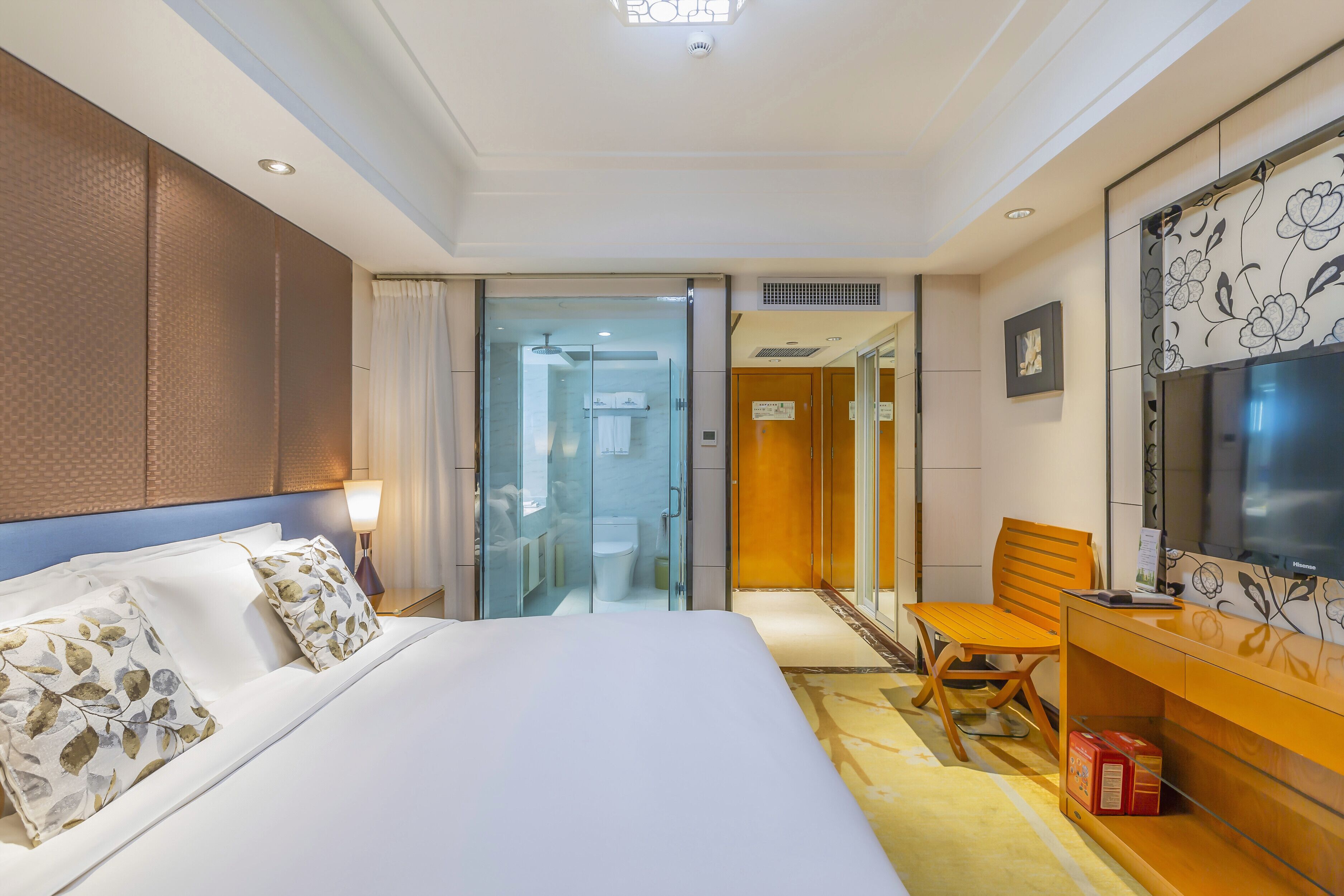 A hotel room with a large bed, a desk with a chair, a television, and a bathroom visible through a glass door.