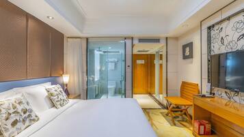 A hotel room with a large bed, a desk with a chair, a television, and a bathroom visible through a glass door.