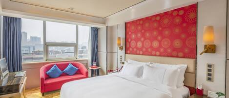 A hotel room with a large bed, a desk with a laptop, a red sofa with blue cushions, a city view, and a red patterned wall.
