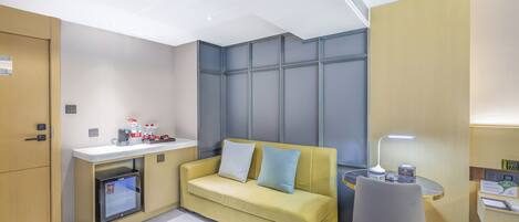 A compact hotel room with a yellow sofa, a small desk with a lamp, a mini-fridge, and a chair.