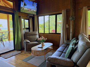 A room with a leather sofa, a wooden coffee table, a chair, a small table with a vase of flowers, and a view of the outdoors through large windows.