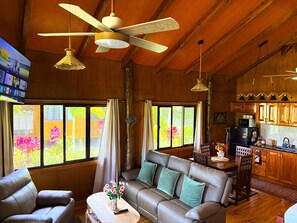 Un salon chaleureux avec un canapé, une table basse et un téléviseur. Le plafond est orné de poutres en bois, et il y a un coin cuisine avec des meubles en bois.
