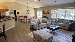 A spacious living area with a large sofa set, a coffee table, and a patterned rug. There is a dining area with a red table and chairs, and a kitchen with a refrigerator and cabinets. - Surfside Hideaway! Dog friendly 3 bed, 2 bath home. Only 1 mile to beach! (Myrtle Beach)