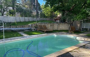 Ein Schwimmbecken mit einer Steinsäule, einer Rasenfläche und einer Holzterrasse mit Liegestühlen.