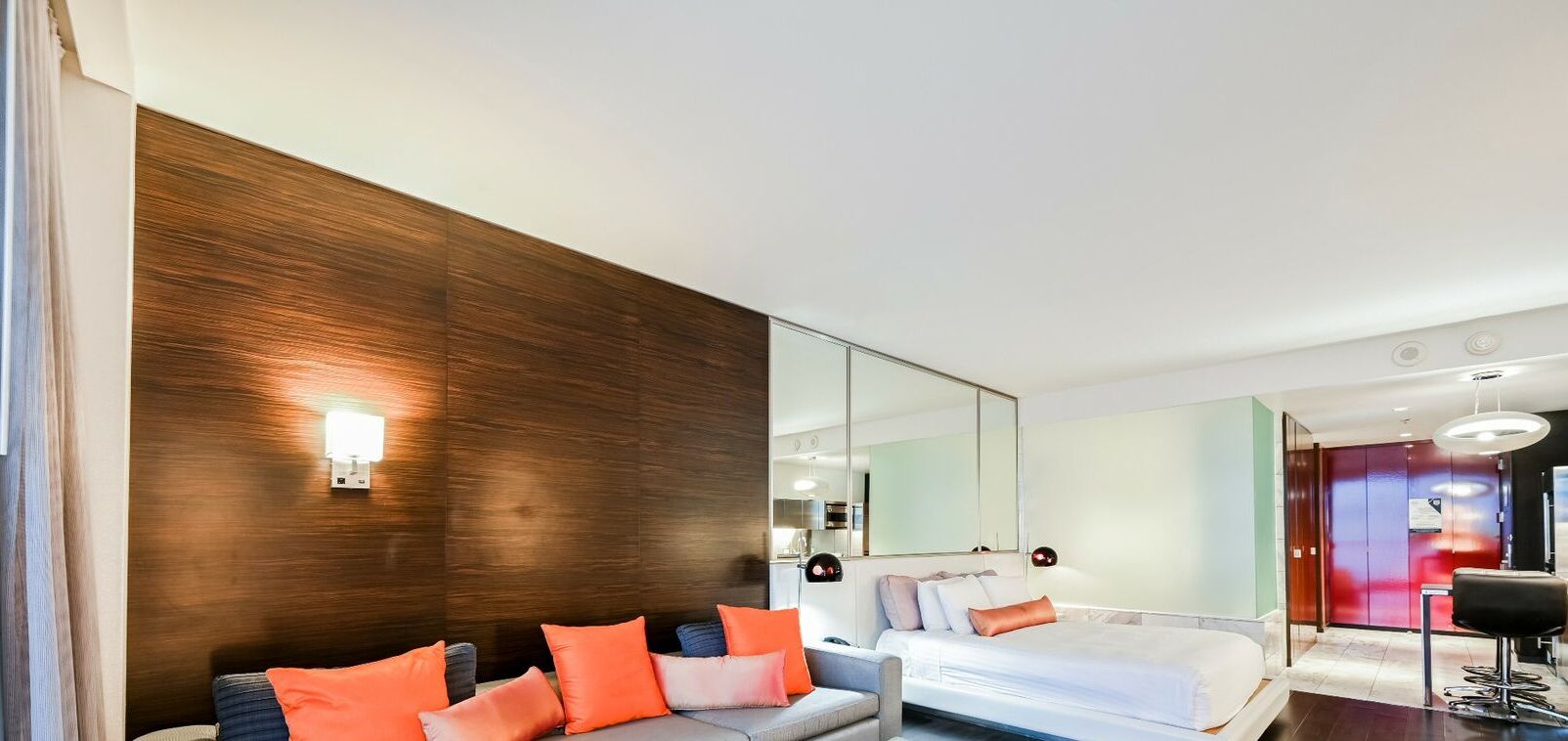 A modern hotel room with a large bed, a sofa with orange cushions, a mirror, and a red door.