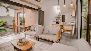 Una sala de estar moderna con sofá, mesa de centro y comedor. Una escalera conduce a otro nivel, y un gran ventanal ofrece vistas del exterior.