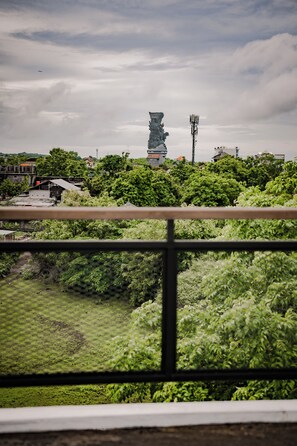 A view from a window with a metal railing, featuring a statue in the distance, surrounded by greenery and buildings.