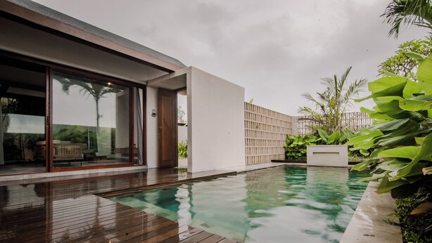 Zona de piscina exterior moderna con cubierta de madera, borde de hormigón y abundante vegetación.