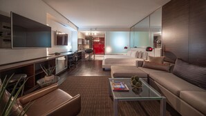 Ein modernes Wohnzimmer mit einer Couch, einem Couchtisch und einem Fernseher. Im Hintergrund befindet sich ein Küchenbereich mit einer roten Tür.