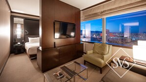 Quarto de hotel com cama, televisão na parede, sofá, mesa de centro e uma grande janela com vista para a cidade.