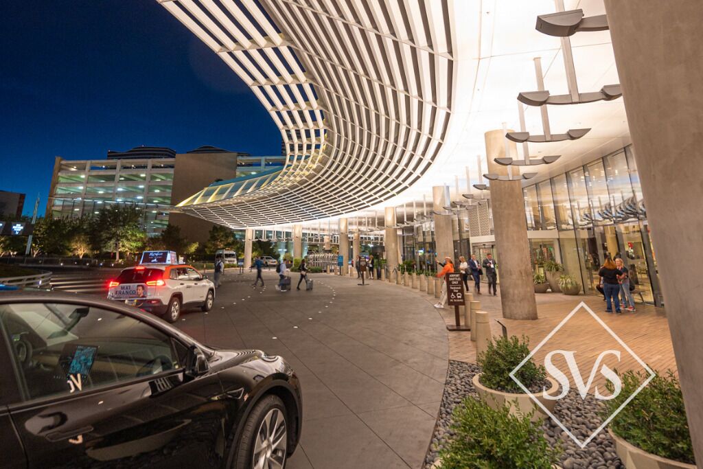 An urban hotel entrance with a modern canopy structure, a paved walkway, and a visible logo.