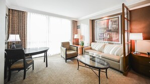 Hotelkamer met een eettafel, twee fauteuils, een bank, een glazen salontafel en een groot raam met gordijnen.