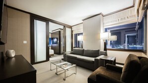 Ein modernes Hotelzimmer mit grauem Sofa, einem Glastisch und Blick auf die beleuchtete Stadt bei Nacht.