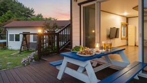 Ein Essbereich im Freien mit einer Holzterrasse, einem Tisch mit Speisen und Biergläsern, und einer Treppe, die zu einem Gebäude führt.
