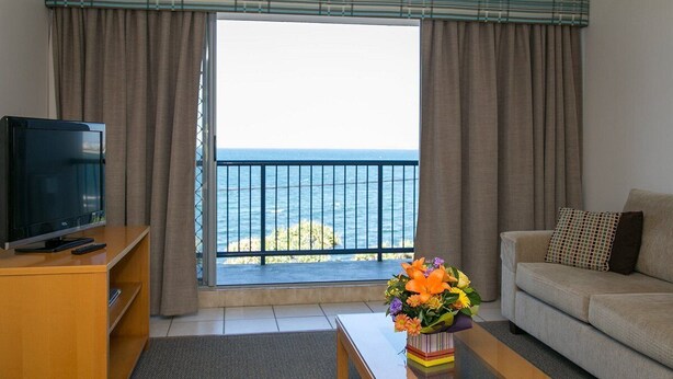 Una stanza con televisione su un mobile in legno, un divano, un tavolino da caffè con un vaso di fiori e vista sul mare da un balcone con tende.