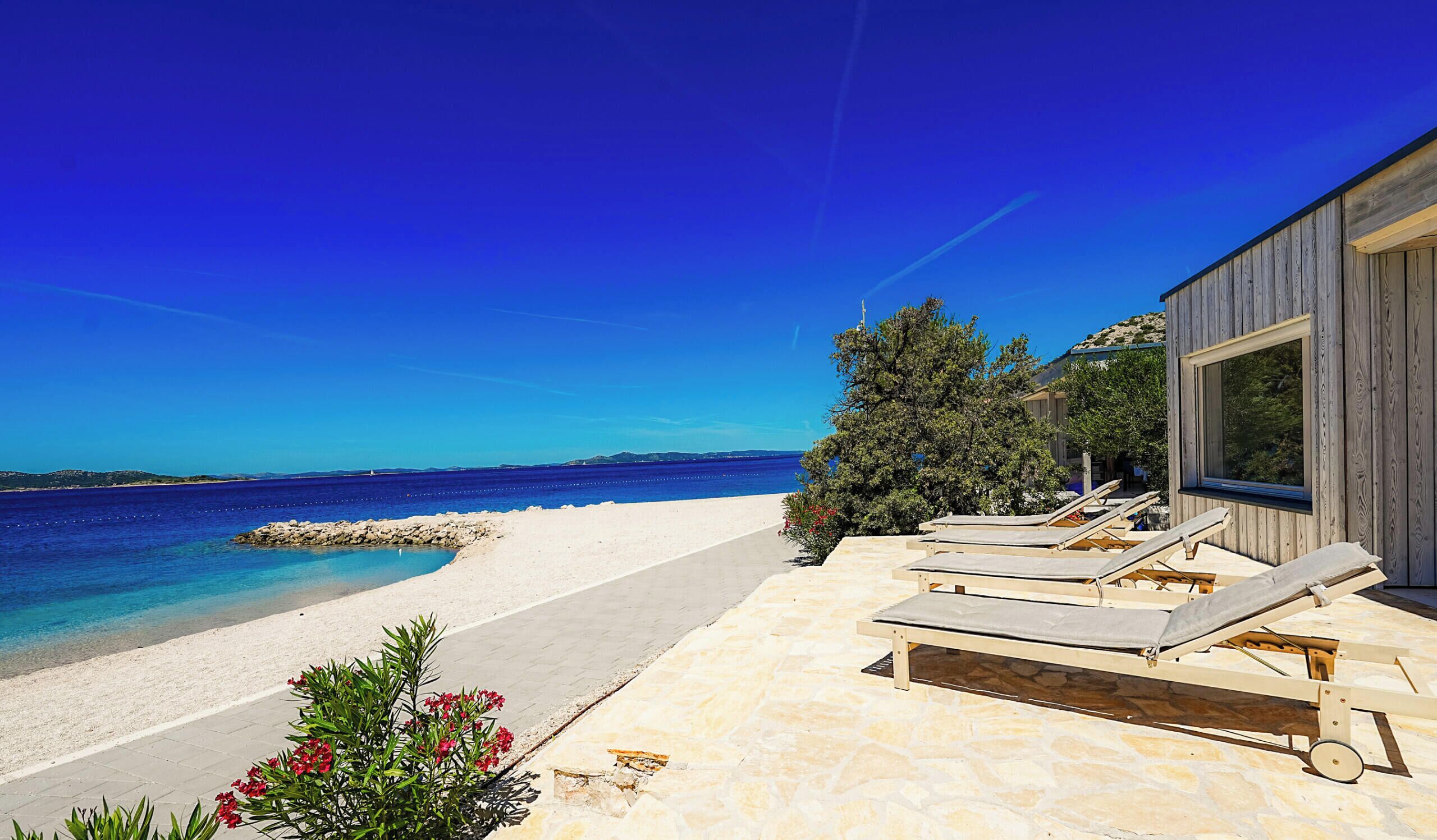 A beachside property with a stone patio, lounge chairs, and a view of the sea.