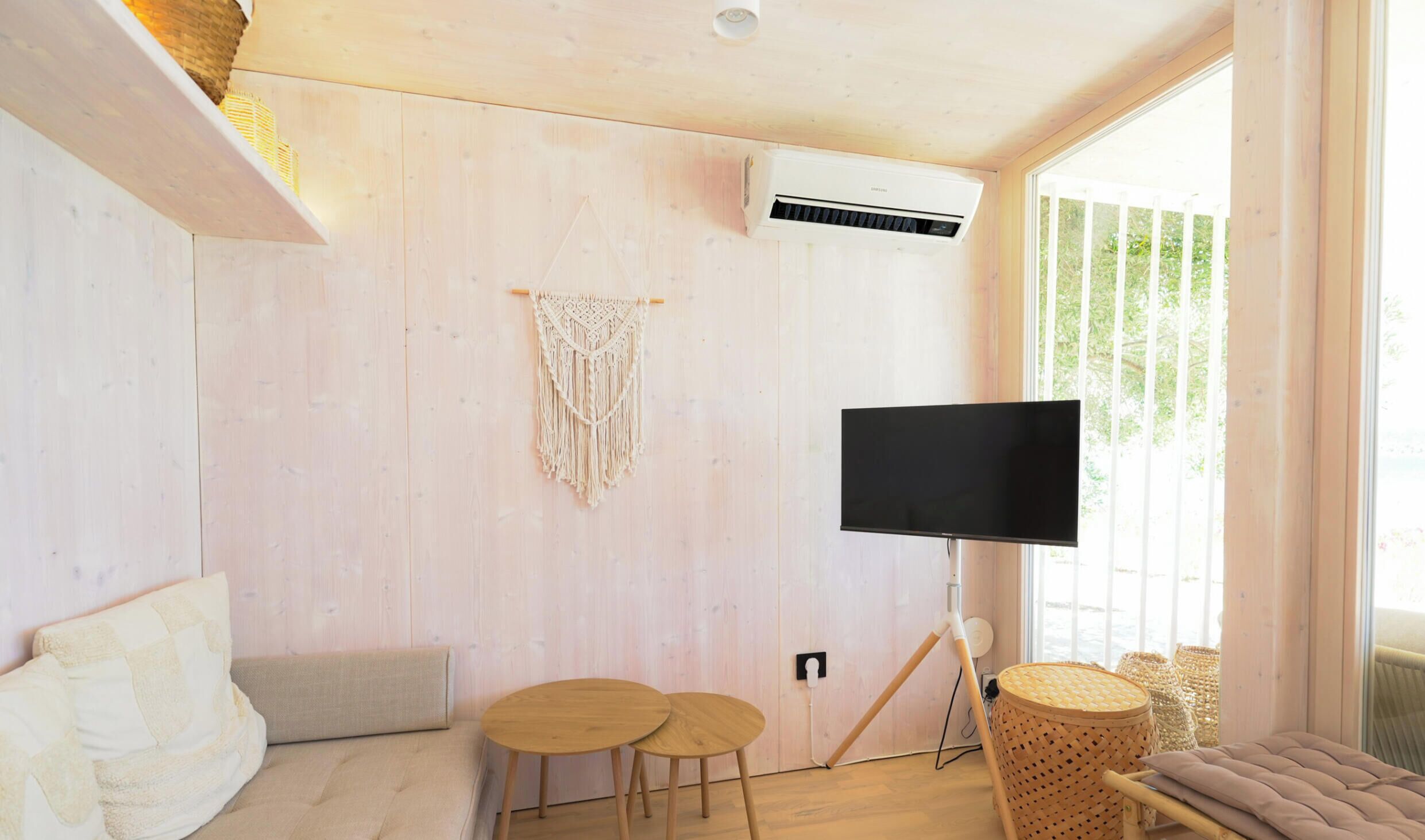 Una stanza con parete in legno, TV a muro, decorazione macramè e un’area salotto con divano e due sgabelli.