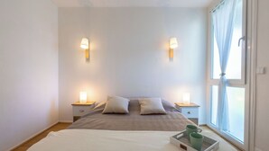 A neatly made bed with a white blanket, two pillows, and a tray with a green mug. There are two bedside tables with lamps, a window with curtains, and a plain wall. - Arcachon Marines by Interhome (Arcachon)