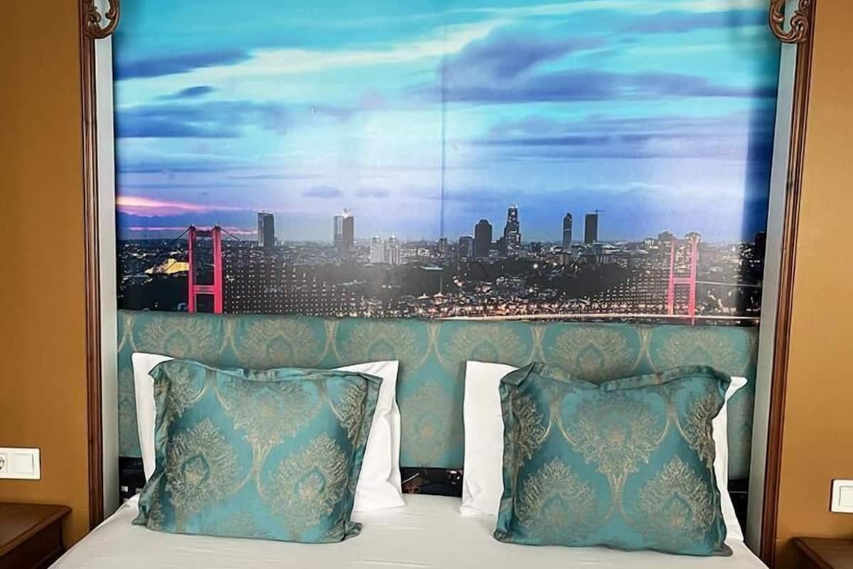A hotel room with a large cityscape mural on the headboard, two teal patterned pillows, and a white bedspread.