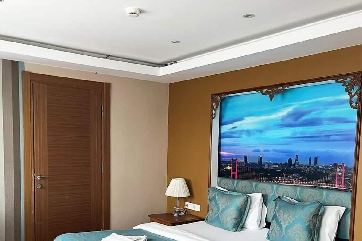 A hotel room with a large bed, a nightstand, a lamp, and a cityscape painting on the wall.