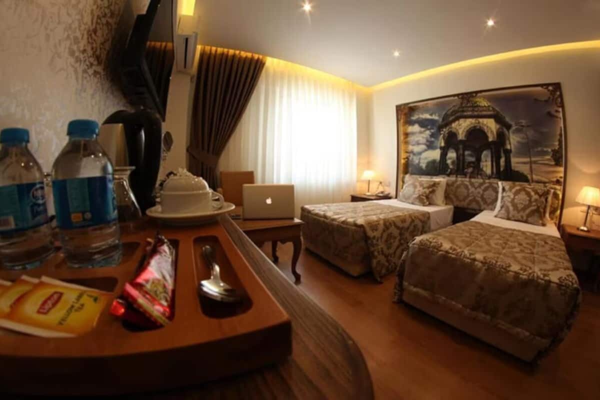 A hotel room with two beds, a desk with a laptop, a chair, a table with water bottles and snacks, and a painting on the wall.