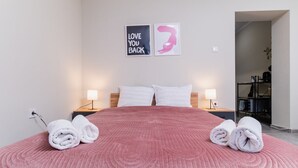A neatly made bed with a pink quilt, two white pillows, and rolled towels on the side. There are two bedside lamps and a wall with two framed pictures. - City Center Studio Apartments 1 (Plovdiv)