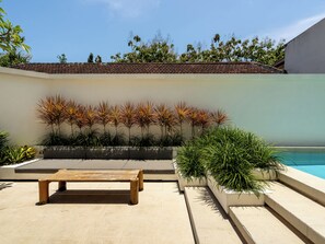 Zona de piscina con un banco de madera, plantas en macetas y vista al mar.