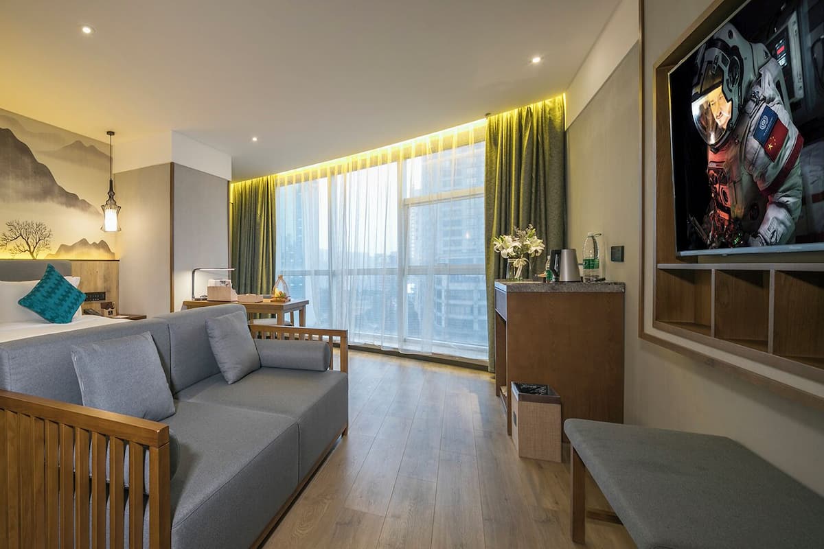 A modern hotel room with a bed, a sofa, a desk, and a large window with curtains.