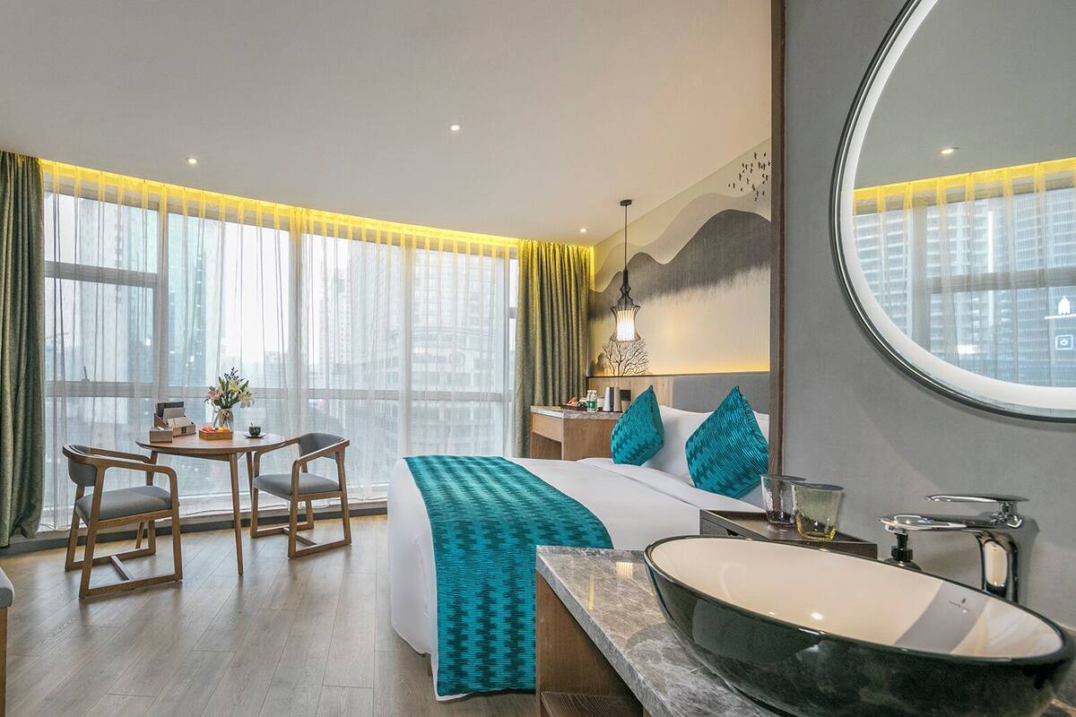 A modern hotel room with a large bed, a round mirror, a vanity with a bathtub, a desk with chairs, and large windows with curtains.