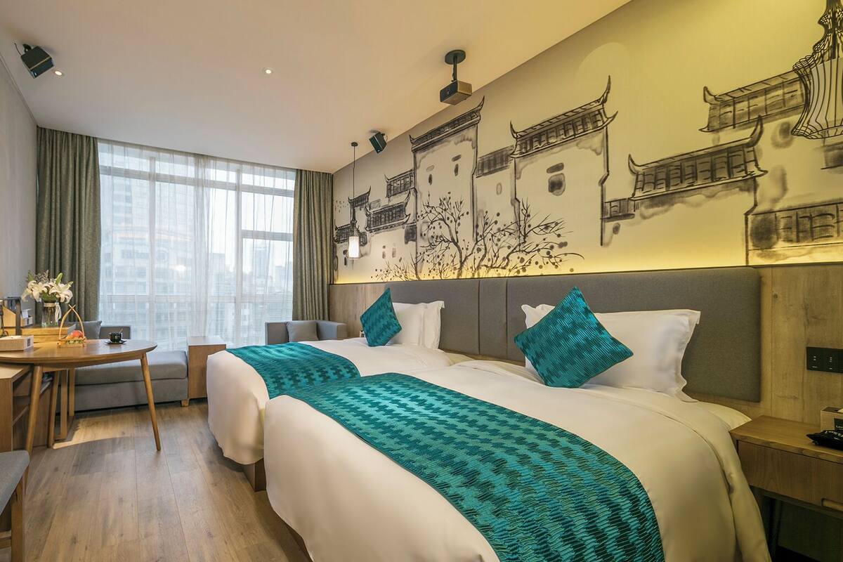 A hotel room with a large bed, two pillows, a bedside table, a desk with a chair, and a large window.
