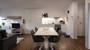 Une cuisine moderne avec des armoires blanches, un four à micro-ondes et une cuisinière. On y trouve une table à manger avec des chaises, ainsi qu’un téléviseur dans l’espace salon.