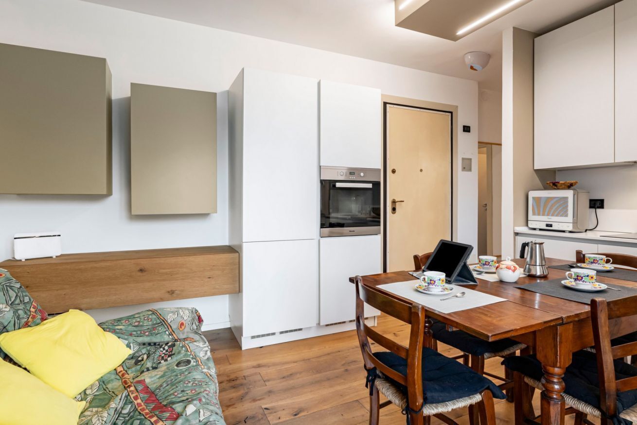Una cucina moderna con mobili bianchi, forno integrato e microonde. C'è un tavolo da pranzo con sedie e un laptop.