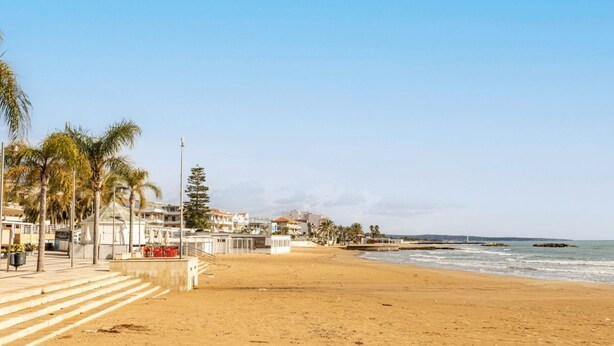 Una playa con escalones que bajan hacia la orilla, palmeras y edificios al fondo.
