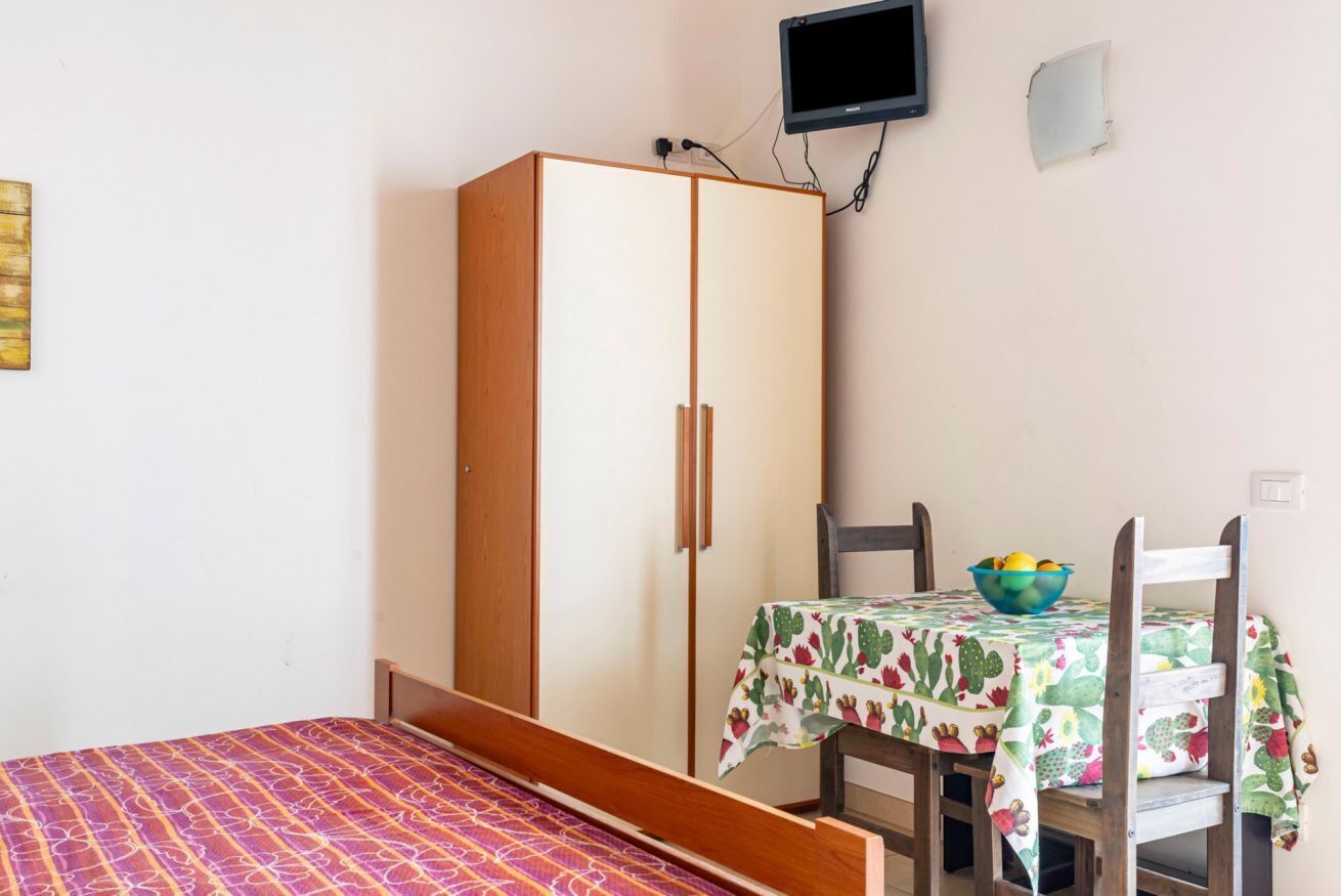 A bedroom with a bed, a wardrobe, a table with a cactus-patterned tablecloth, and a television mounted on the wall.