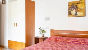 Un dormitorio con cama, armario de madera, mesita de noche con una planta y una obra de arte colgada en la pared.