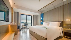 A modern hotel room with a large bed, a desk, and a chair. There is a view of the city through the window.