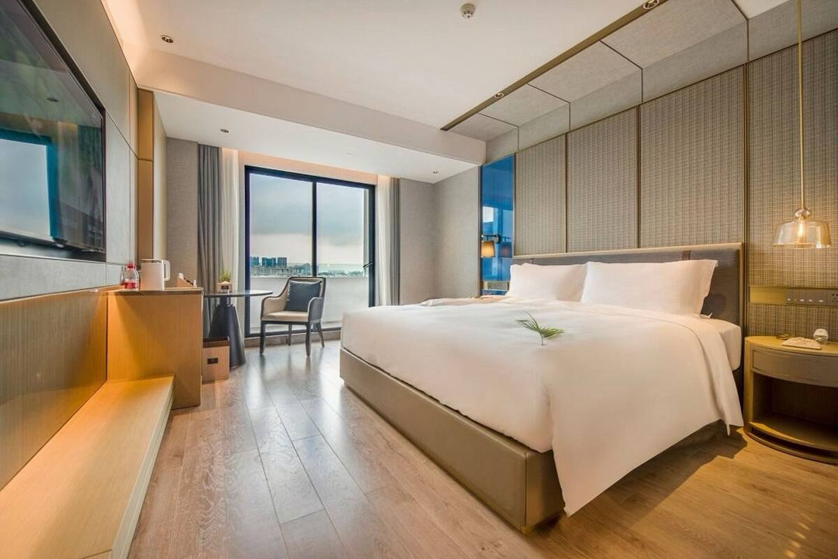 Une chambre d’hôtel moderne avec un grand lit, un bureau et une chaise. La fenêtre offre une vue sur la ville.