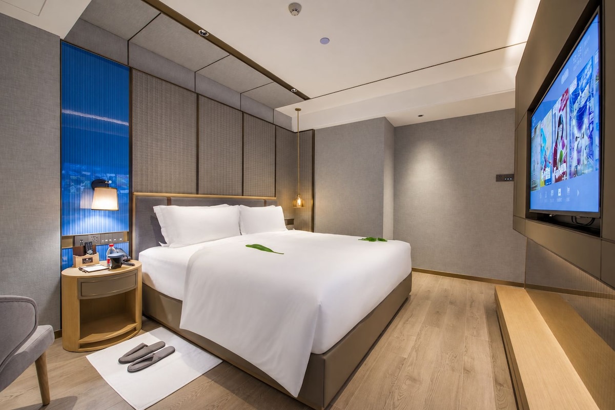 Et moderne hotelværelse med en stor seng, et fladskærms-tv, et sengebord med lampe og udsigt over bybilledet gennem vinduet.
