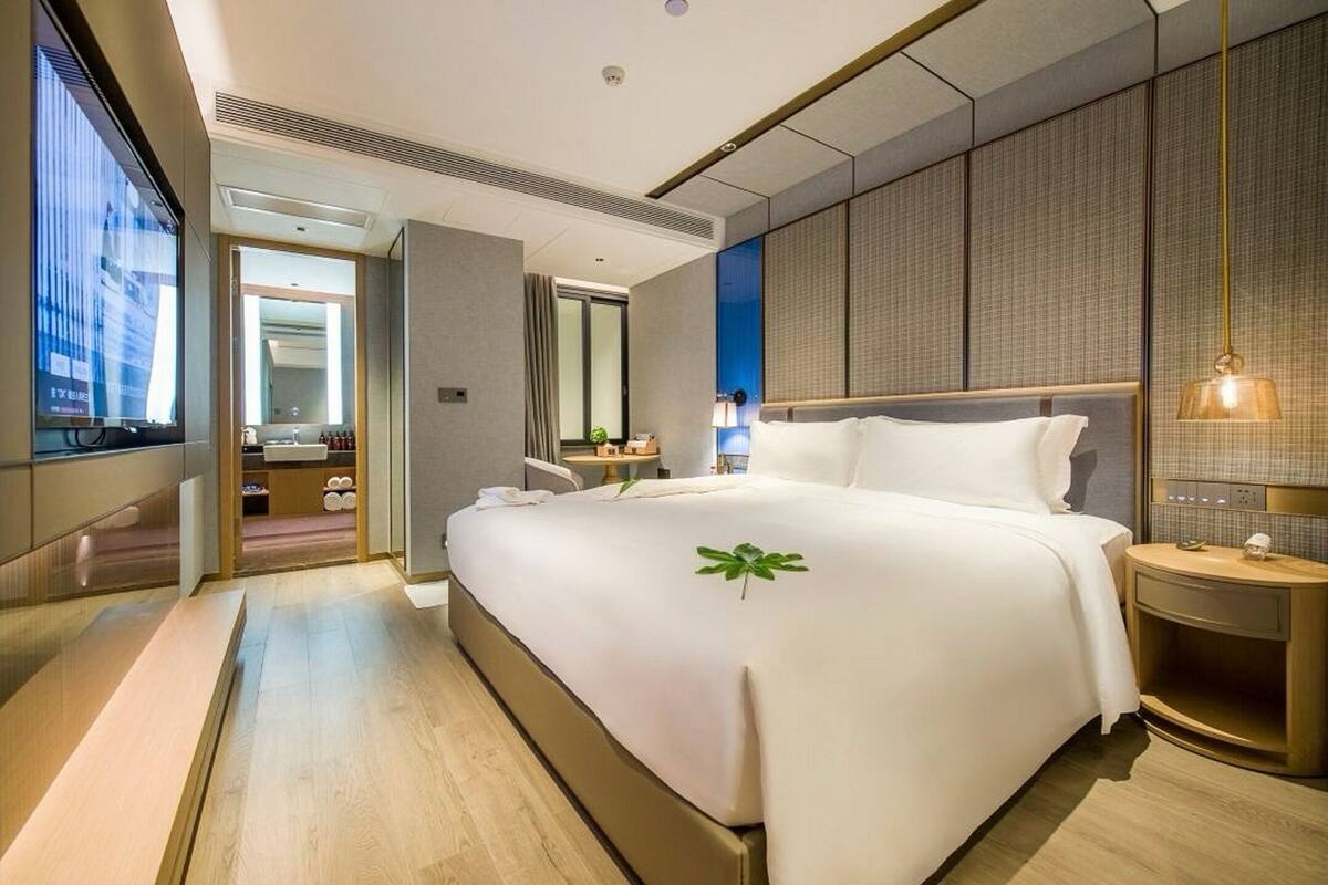 Une chambre d’hôtel moderne avec un grand lit, une table de chevet, une commode, une fenêtre et une salle de bain visible à travers une porte ouverte.