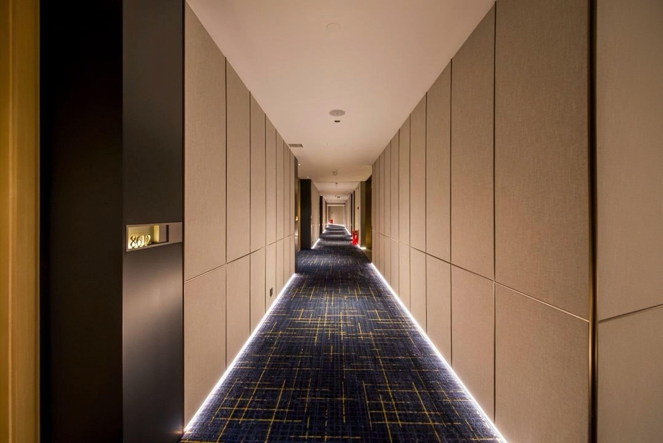 Een lange, smalle hotelgang met een patroon tapijt en beige wanden.