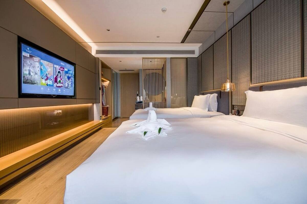 Une chambre d’hôtel moderne dotée d’un grand téléviseur à écran plat fixé au mur, d’un lit bien fait avec du linge de maison blanc et d’un banc en bois le long du mur.
