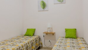 Un petit logement composé d’une seule pièce, avec deux lits, chacun recouvert d’une couvre-lit à motifs et agrémenté de coussins verts. Une lampe fixée au mur est posée sur une étagère, et deux feuilles encadrées ornent le mur.